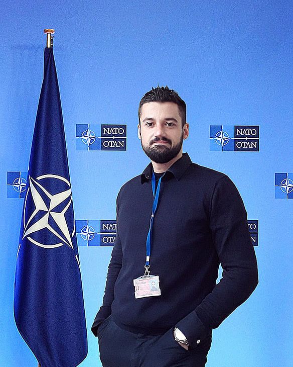 George Alex Stelea stands in front of a blue backdrop featuring repeated NATO/OTAN logos. He is wearing a dark long-sleeve top and a lanyard with an ID badge, with one hand in his pocket. A NATO flag is positioned to his left. He is facing the camera with a neutral, confident expression.